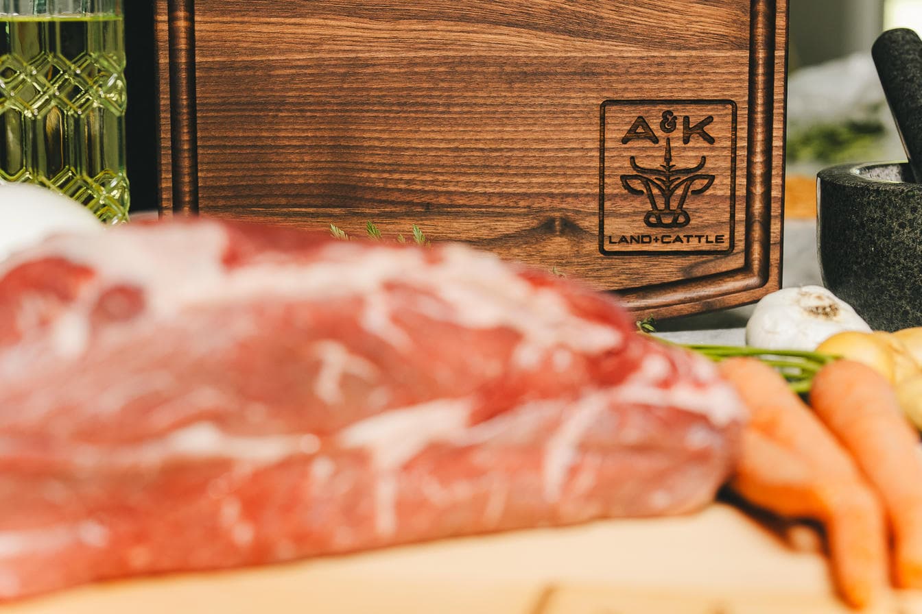 Raw meat on a wooden cutting board with a brand logo, surrounded by vegetables.