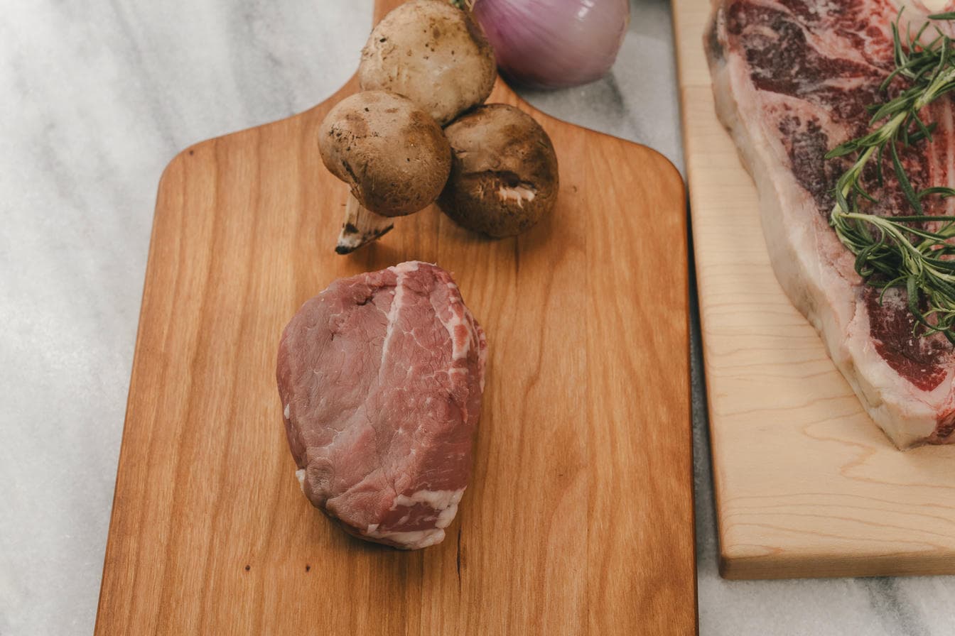 Raw meat, mushrooms, and a red onion on a wooden cutting board with a marble background.