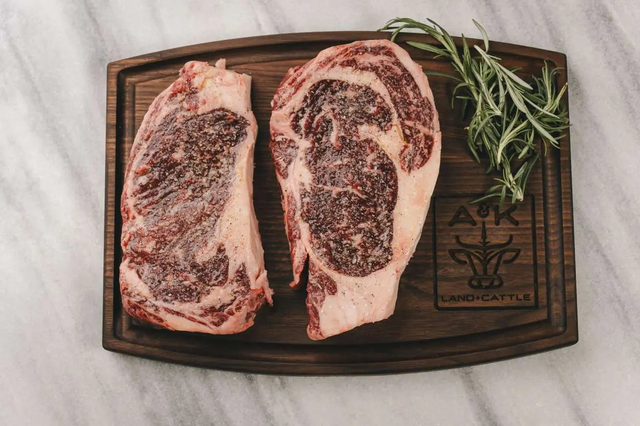 Two raw steaks on a wooden cutting board with rosemary, branded 'A&K Ranch Cattle'.