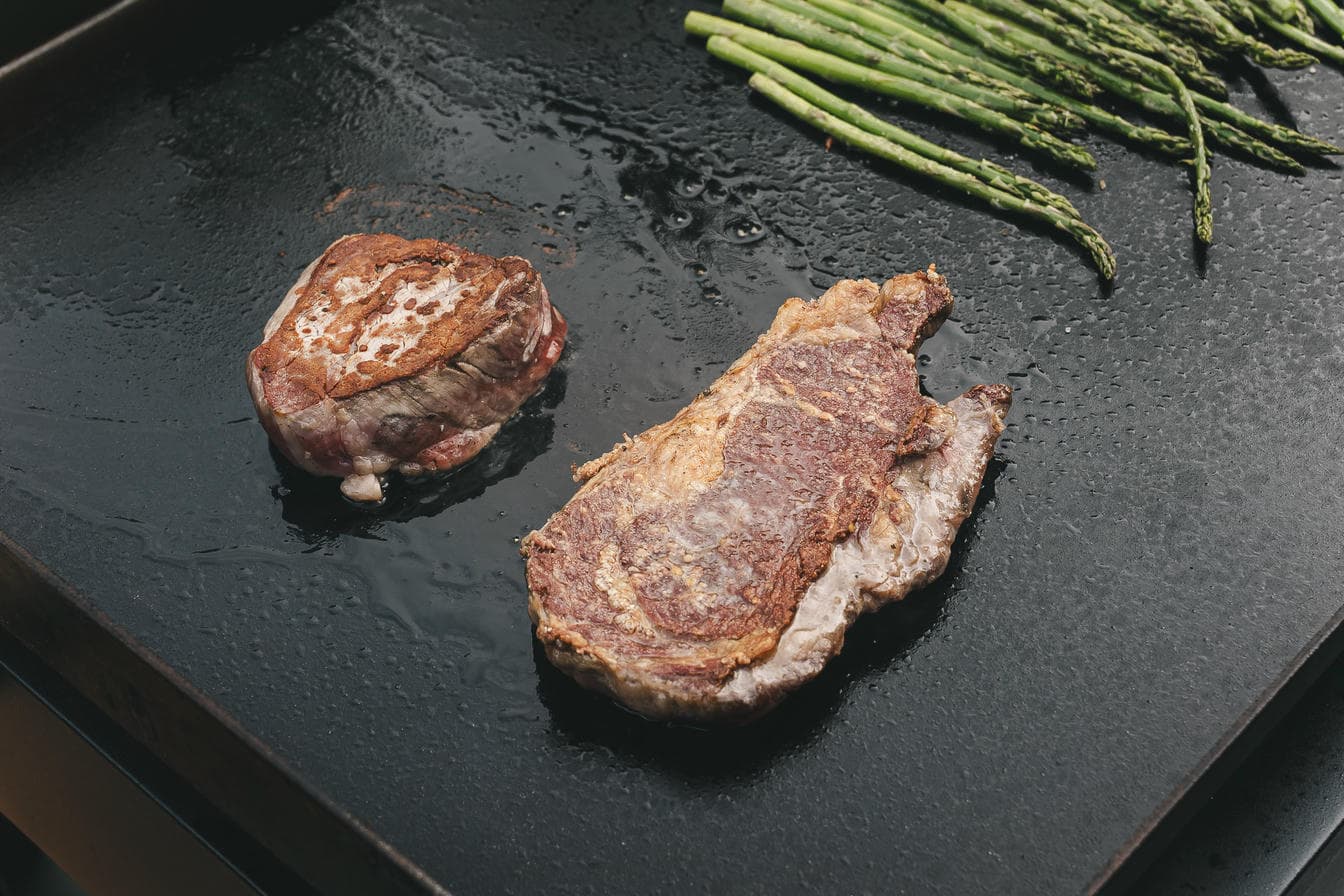 Two seared A&K dry aged steaks on a hot griddle with asparagus nearby
