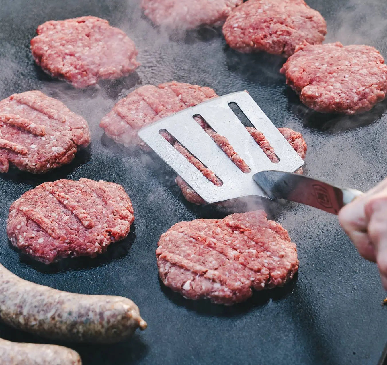 Raw hamburger patties being cooked on a grill with a spatula.