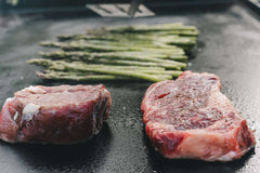 Two raw steaks and asparagus on a grill pan.