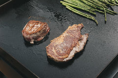 Two seared A&K dry aged steaks on a hot griddle with asparagus nearby